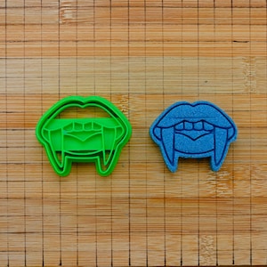 May include: A green plastic vampire mouth cookie cutter and a blue vampire mouth cookie stamp. The green cutter features an open mouth with sharp fangs and visible teeth, while the blue stamp creates a matching impression of the same design.