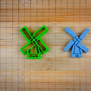 Puede incluir: Dos cortadores de galletas de plástico con forma de molinos de viento. Uno es verde y el otro es azul. Están sobre una tabla de cortar de madera.
