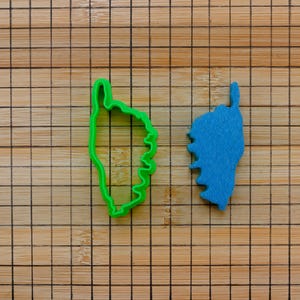 Può includere: Due formine per biscotti a forma di Corsica. Uno è verde brillante, l'altro blu. Sono posizionati su un tagliere di legno con un motivo a griglia.