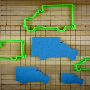 May include: Five cookie cutters shaped like delivery vans. Two are green and three are blue. The cutters are on a wooden chopping board with a grid pattern.