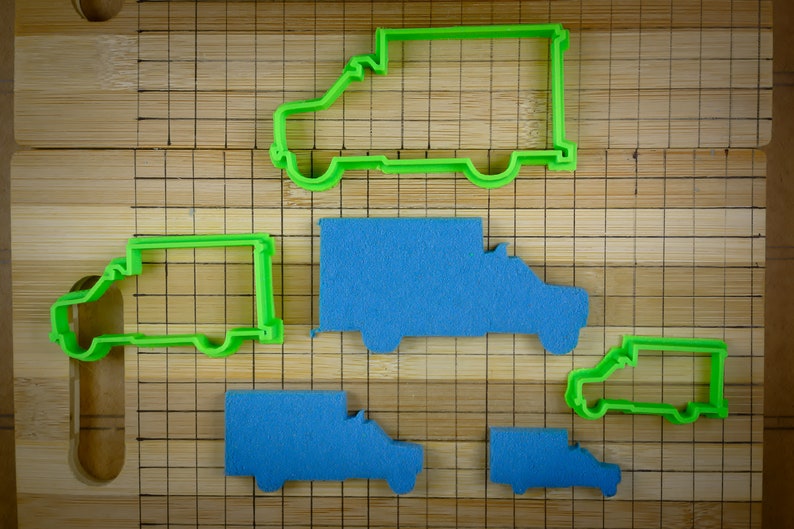 May include: Five cookie cutters shaped like delivery vans. Two are green and three are blue. The cutters are on a wooden chopping board with a grid pattern.