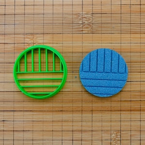 Könnte beinhalten: Ein grüner, kreisförmiger Ausstecher mit geschlitztem Design liegt neben einem blauen, kreisförmigen, strukturierten Objekt. Die Objekte befinden sich auf einem Bambus-Schneidebrett.