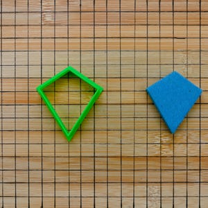 Può includere: Un tagliapasta di plastica verde a forma di diamante e un tagliapasta di feltro blu a forma di diamante su un tagliere di legno con un motivo a griglia.