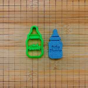 May include: Two baby bottle-shaped cookie cutters. One is green and says "Baby Shower" in reverse. The other is blue and says "Baby Shower". Both are on a wooden cutting board.