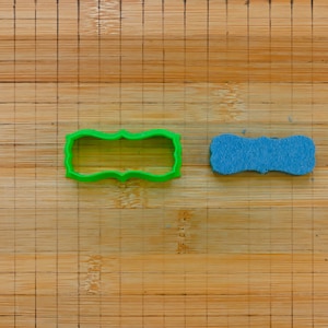 Puede incluir: Un cortador de galletas de plástico verde en forma de rectángulo con esquinas redondeadas y una esponja de espuma azul sobre una tabla de cortar de madera.