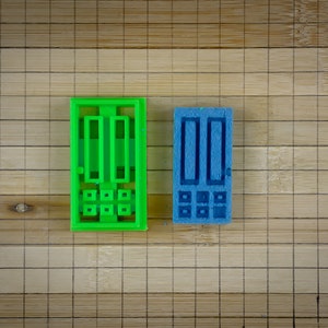 May include: Two plastic molds for making chocolate, one green and one blue. Each mold has a rectangular shape with three vertical lines and three horizontal lines, creating a grid pattern.