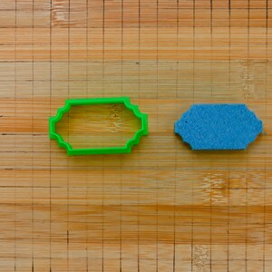 May include: Two cookie cutters on a wooden surface. The cookie cutter on the left is green and has a scalloped edge. The cookie cutter on the right is blue and has a smooth edge.