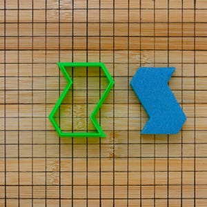 May include: Two plastic cookie cutters, one green and one blue, shaped like a chevron or zigzag. The cutters are on a wooden cutting board with a grid pattern.