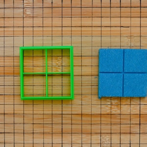 May include: A green square cookie cutter with a grid design and a blue square divided into four sections, placed on a wooden surface with a grid pattern.