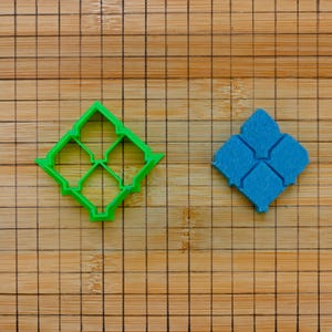 Puede incluir: Un cortador de galletas de plástico verde en forma de diamante con un diseño floral calado, junto a un objeto azul en forma de diamante con un diseño similar. Los objetos están sobre una tabla de cortar de madera.