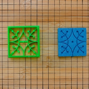 Op de afbeelding: Een groene vierkante koekjesvorm met een bloemenpatroon naast een blauwe vierkant met hetzelfde bloemenpatroon. De objecten liggen op een houten snijplank met een rasterpatroon.
