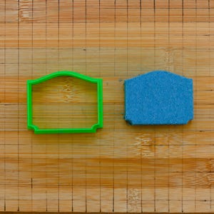 Könnte beinhalten: Ein grüner Ausstecher in Form eines Schildes neben einem blauen Schwamm. Die Objekte liegen auf einem hellbraunen Bambus-Schneidebrett. Der Ausstecher hat einen dekorativen Rand.