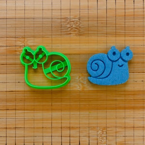 May include: Two snail-shaped items on a wooden chopping board. One is a bright green plastic cookie cutter, and the other is a blue felt snail. The cookie cutter has a detailed outline, while the felt snail has a simple design.