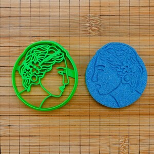 May include: A bright green circular cookie cutter featuring a profile of a person with wavy hair and a flower accessory, alongside a blue circular embossed impression of the same design, showing the detailed facial features and hair texture.