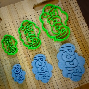 May include: Five cookie cutters on a wooden surface. The cookie cutters are green and blue and shaped like clouds with words on them. The words on the cookie cutters are "Foodie", "Foodie", "Foodie", "Roodie", and "Roodie".