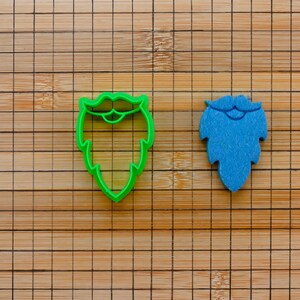May include: Two cookie cutters, one green and one blue, shaped like beards. The green beard has a mustache.