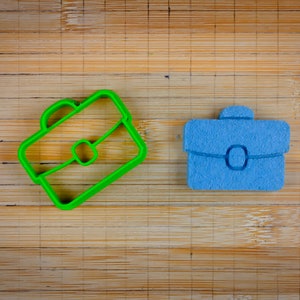May include: A green plastic briefcase-shaped cookie cutter and a blue briefcase-shaped object sit on a bamboo surface. The cookie cutter is a bright green outline, while the blue object has a textured appearance.