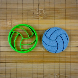 Könnte beinhalten: Ein grüner Plastik-Volleyball-Ausstecher und ein blauer, volleyballförmiger Keks auf einer Holzoberfläche.