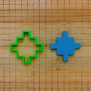 Pode incluir: Um cortador de biscoitos de plástico verde em forma de cruz posicionado ao lado de uma peça em forma de cruz feita de material azul texturizado. Ambos os itens estão sobre uma superfície de madeira castanho-clara com uma grade escura.