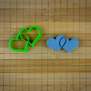 Pode incluir: Dois cortadores de biscoitos em forma de coração verde e dois corações de feltro azul em uma tábua de cortar de madeira.