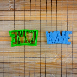 Puede incluir: Dos cortadores de galletas, uno verde y otro azul, con forma de las palabras "Te amo" con un corazón en el medio de cada palabra.