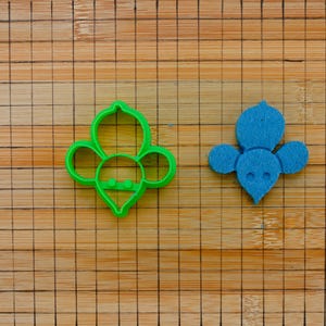 Può includere: Due oggetti a forma di ape su una superficie di legno. Uno è un tagliabiscotti in plastica verde brillante, l'altro è un ritaglio di feltro blu. Le forme delle api hanno ali arrotondate e un fondo a punta.