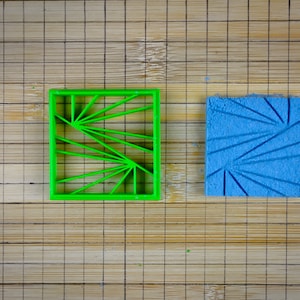 May include: A green plastic square cookie cutter with a geometric design next to a blue square with the same pattern. The objects are on a bamboo cutting board.