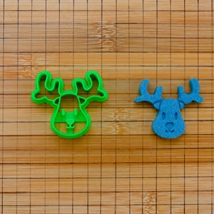 Può includere: Due oggetti a forma di alce su una superficie di legno. Uno è un tagliapasta in plastica verde brillante, l'altro è una piccola decorazione in feltro blu. Le forme di alce hanno corna e lineamenti del viso dettagliati.