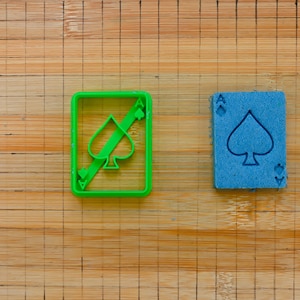 May include: Two plastic cookie cutters shaped like playing cards. The cutter on the left is green and features an ace of spades with a diagonal line through it. The cutter on the right is blue and features an ace of spades.