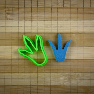 Op de afbeelding: Twee dinosaurus-voetafdruk koekjesvormen, een groene en een blauwe, op een houten oppervlak.