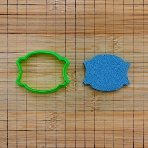 May include: A green plastic cookie cutter and a blue textured object sit on a wooden surface. The cookie cutter is in the shape of a decorative shield. The blue object is also shield-shaped.