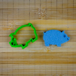 May include: A green plastic pig cookie cutter and a blue felt pig on a wooden cutting board.