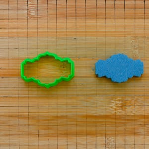 May include: Two cookie cutters on a wooden surface. The cutter on the left is green and shaped like a banner. The cutter on the right is blue and shaped like a cloud.