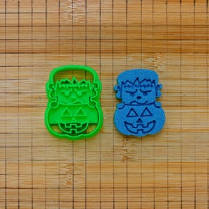 Può includere: Un tagliabiscotti in plastica verde brillante a forma di testa di mostro di Frankenstein sopra una zucca, posizionato accanto a un'impronta testurizzata blu abbinata dello stesso design, entrambi con tratti del viso e forme geometriche.