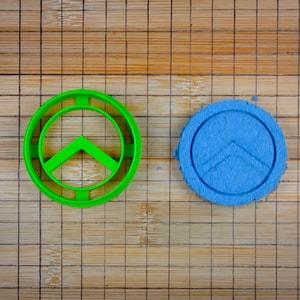 Può includere: Un tagliapasta di plastica verde a forma di scudo spartano, accanto a un biscotto blu con lo stesso design.