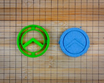 Könnte beinhalten: Ein grüner Plastik-Ausstecher in Form eines spartanischen Schildes, neben einem blauen Keks mit dem gleichen Design.