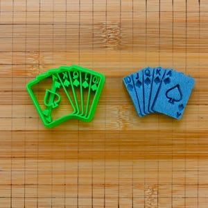 Puede incluir: Dos juegos de cortadores de galletas con forma de cartas de juego, uno verde y otro azul. El juego verde es una escalera real de picas, y el juego azul es una escalera real de tréboles.