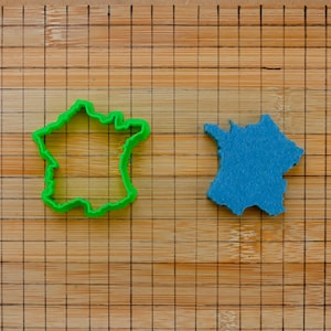 May include: A green plastic cookie cutter and a blue felt cutout, both shaped like France, are on a bamboo cutting board. The board has a grid pattern. The cookie cutter is a vibrant green.