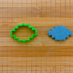 May include: A green plastic cookie cutter shaped like a rounded rectangle with a decorative edge and a blue felt cookie cutout on a wooden cutting board.