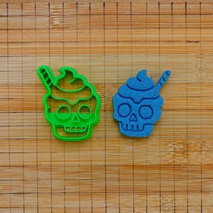Könnte beinhalten: Ein hellgrüner Kunststoff-Ausstecher in Form eines Totenkopfes mit einer Sahnehaube und einem Strohhalm, platziert neben einem passenden blauen, texturierten Totenkopf-Ausschnitt aus körnigem Material auf einer Holzoberfläche mit Gitter.