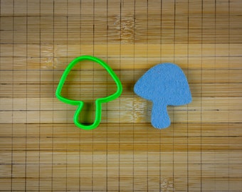 Peut inclure: Un emporte-pièce en forme de champignon en plastique vert et un champignon en feutre bleu sur une planche à découper en bambou.