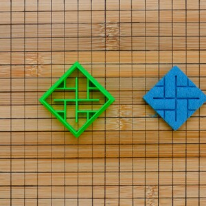 Puede incluir: Dos objetos en forma de diamante sobre una superficie de madera. Uno es un diseño verde brillante de marco abierto. El otro es un cuadrado azul sólido con un patrón geométrico en relieve. La superficie de madera tiene un patrón de cuadrícula.