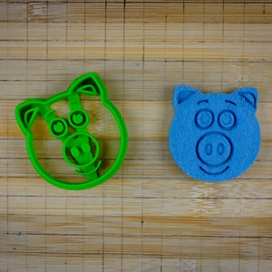 May include: Two pig-shaped cookie cutters, one green and one blue, on a bamboo surface.