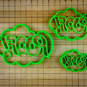May include: Three green plastic cookie cutters shaped like a speech bubble with the word "FOOD" inside. The cutters are on a wooden surface.