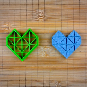 May include: Two heart-shaped cookie cutters, one green and one blue, with a geometric design. The cutters are on a bamboo cutting board.