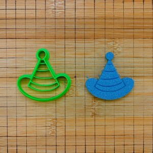 May include: A green plastic party hat cookie cutter and a blue textured party hat shape. The party hat design features a pointed top with a small circular pom-pom detail and a wide brim, accented by three horizontal lines across the cone section.