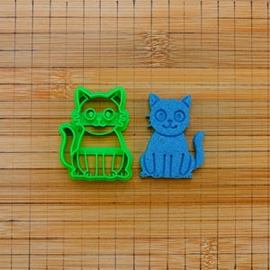 May include: A bright green cat-shaped cookie cutter positioned next to a solid blue cat-shaped cutout. The green cutter features an open frame design with detailed facial features and whiskers, while the blue cutout displays a matching cat silhouette.