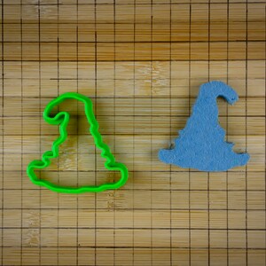 Pode incluir: Dois cortadores de biscoitos em forma de chapéu de bruxa, um verde e outro azul, em uma tábua de cortar de bambu.