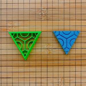 May include: Two triangular shaped cookie cutters, one green and one blue, with geometric patterns. The cutters are on a wooden surface.