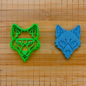 Puede incluir: Dos cortadores de galletas con forma de cabeza de lobo, uno verde y otro azul, sobre una superficie de madera.